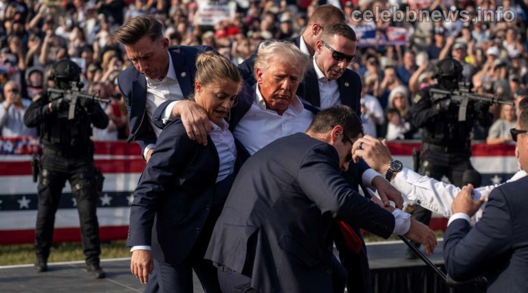 Security personnel assist a man in a light blue shirt surrounded by officials at a rally with crowds in background